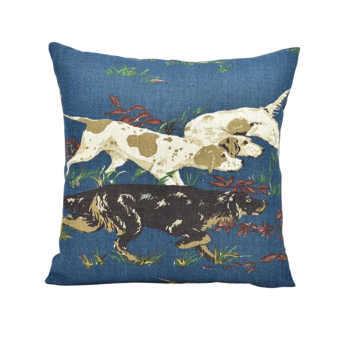 A blue throw pillow with a pattern of three dogs, one brown and two white, walking through a grassy field with leaves on the ground.